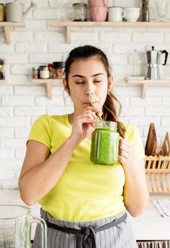 Healthy eating, dieting concept. Young brunette woman drinking green smoothie at home kitchen from mason jar