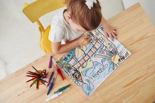 Sitting on the yellow chair. Cute little girl in art school draws her first paintings by pencils and markers.