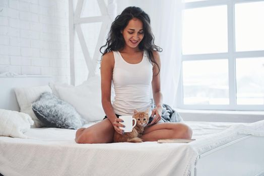 Nice morning. Cute brunette sitting on the white bed with cup of coffee and smartphone in hand.