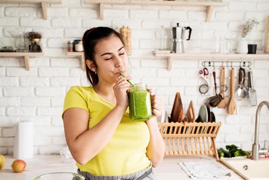 Healthy eating, dieting concept. Young brunette woman drinking green smoothie at home kitchen from mason jar