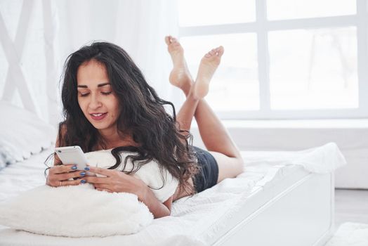 Conversation with friends using mobile device. Brunette having a rest on the white couch in her bedroom at daytime.