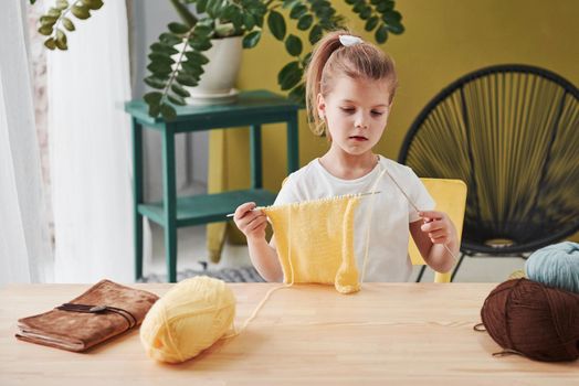 Concentrated at process. Kid is knitting at home. Cute little girl sitting near the wooden table is learning some new stuff.