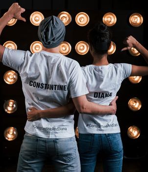 Shows thumbs on themselves. Back view of couple in shirts with their names on it standing next to studio lights.