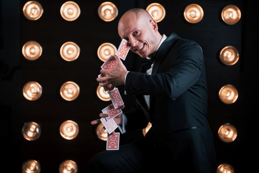 Smiling during the process. Magician in black suit and with playing cards standing in the room with special lighting at backstage.