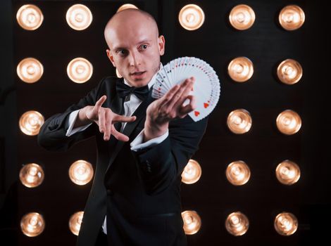 Dexterity of hands. Magician in black suit and with playing cards standing in the room with special lighting at backstage.