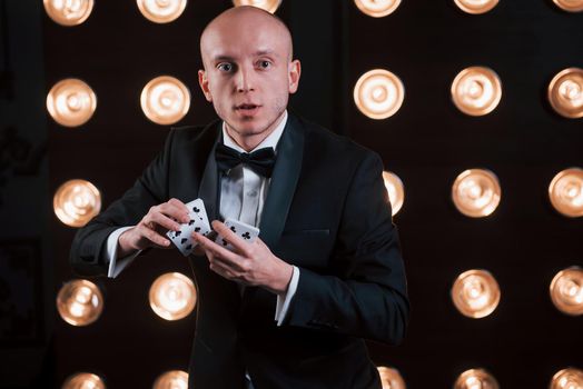 Shuffles the cards. Magician in black suit standing in the room with special lighting at backstage.