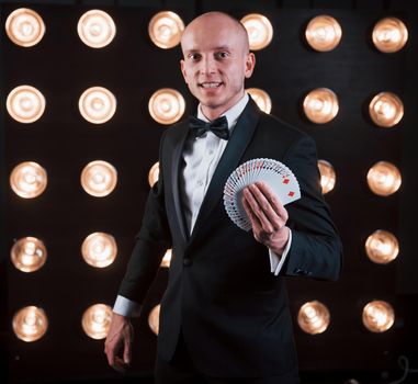 Smiling while showing deck of cards. Magician in black suit standing in the room with special lighting at backstage.