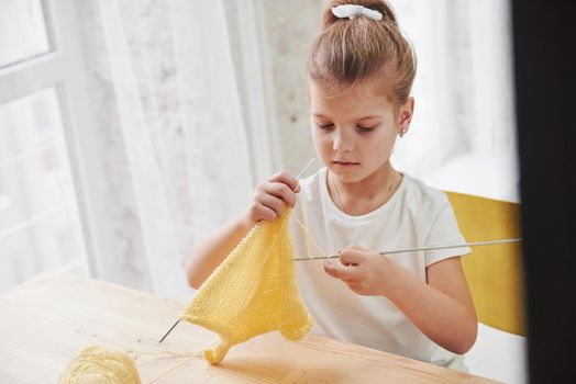 Daylight coming from the window. Kid is knitting at home. Cute little girl sitting near the wooden table is learning some new stuff.