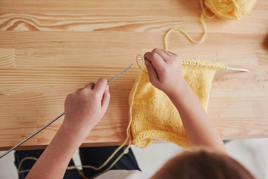 Top view. Kid is knitting at home. Cute little girl sitting near the wooden table is learning some new stuff.