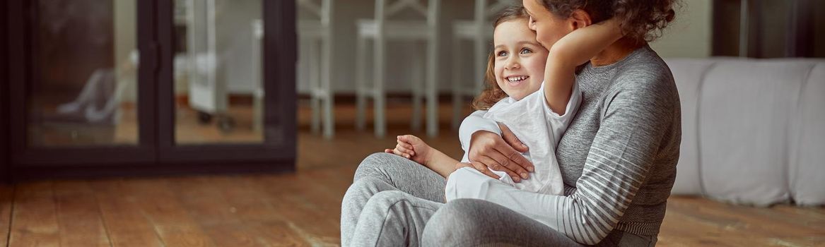 Cheerful woman is cuddling with girl while doing gymnastics on mat in cozy living room