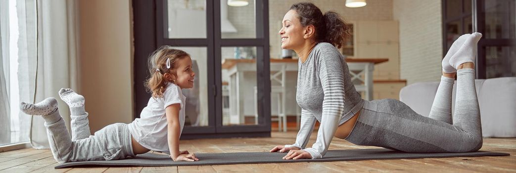 Low angle of cheerful woman doing exercises together with cute girl on floor for improving flexibility