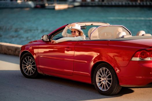 Outdoor summer portrait of stylish blonde woman driving red car convertible. Fashionable attractive woman with blond hair in a white hat in a red car. Sunny bright colors taken outdoors against the sea