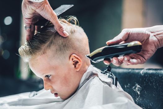 Lifestyle photo. Barber shop little blond boy customer. Hairdresser man work move hand comb tool scissors trimmer white cape. Modern style kid short hair cut. Fashion business service for child.