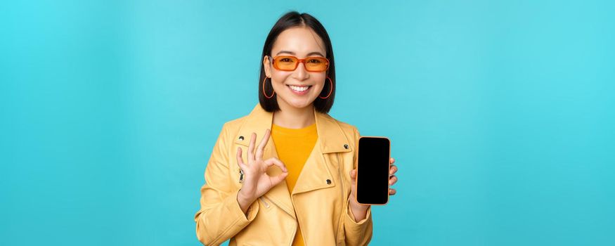 Smiling korean woman showing mobile phone app interface, smartphone application, recommending on cellphone, standing over blue background.