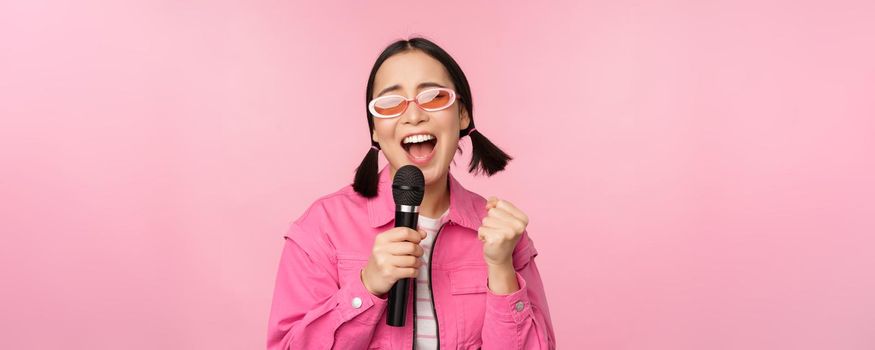 Happy beautiful asian girl singing with mic, using microphone, enjoying karaoke, posing against pink studio background.