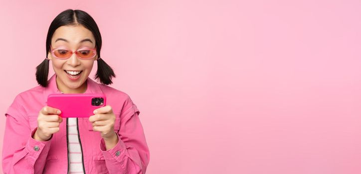 Portrait of happy asian girl playing on smartphone, watching videos on mobile phone app, standing over pink background.