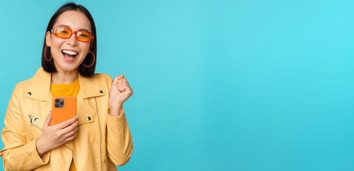 Enthusiastic smiling asian girl in sunglasses, holding mobile phone and dancing, laughing with smartphone, standing over blue background.