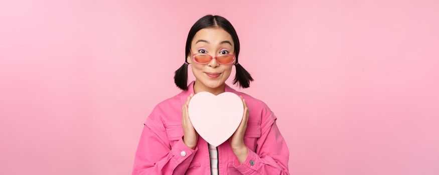 Beautiful asian girl smiling happy, showing heart gift box and looking excited at camera, standing over pink romantic background.