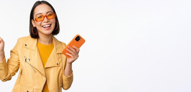 Image of stylish korean girl dancing with smartphone, laughing happy and smiling, standing over white background. Copy space