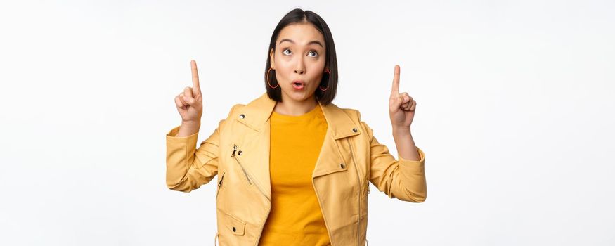 Image of smiling asian brunette woman pointing fingers up, showing advertisement with happy face, posing against white background.