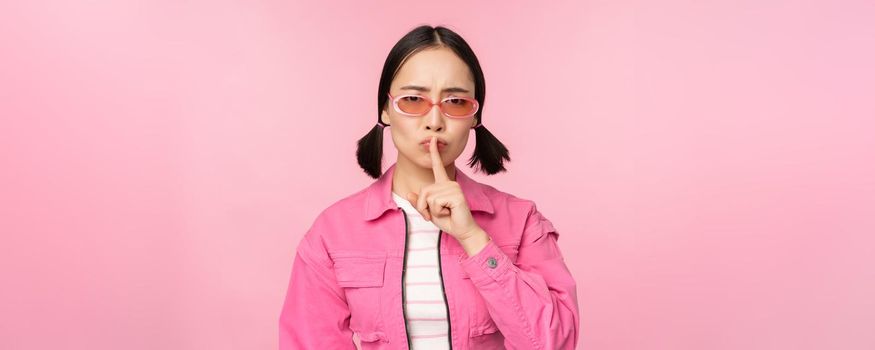 Modern korean girl in stylish spring outfit, sunglasses, showing shush, hush sign, press finger to lips, taboo gesture, standing over pink background.