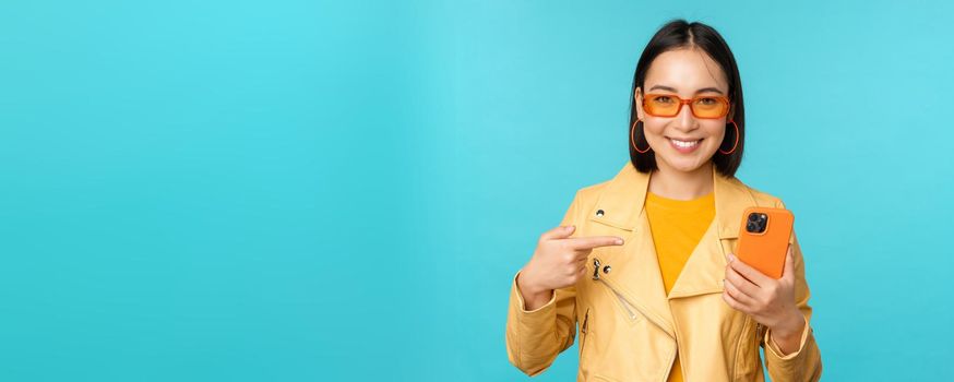 Young stylish asian female model in trendy sunglasses, spring outfit, showing mobile phone, pointing at smartphone and smiling, standing over blue background.
