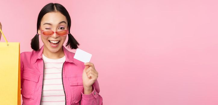 Happy young asian woman showing credit card for shopping, holding bag, buying on sale, going to the shop, store, standing over pink background.