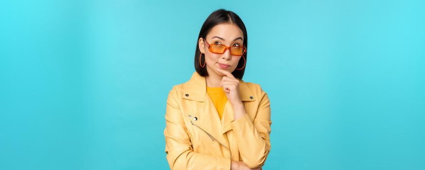 Portrait of asian woman thinking, looking thoughtful, searching ideas or solution, wearing sunglasses, standing over blue background.