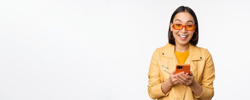 Modern asian girl in sunglasses using her mobile phone, smiling and looking happy, posing against white background. Copy space