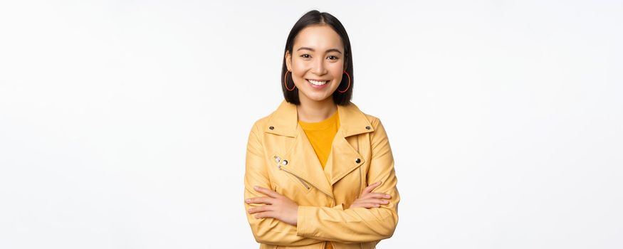 Portrait of asian woman in yellow jacket, smiling and looking happy, standing over white background. Copy space