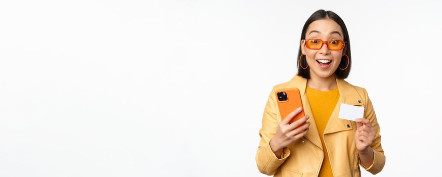 Online shopping. Stylish asian female model in sunglasses, holding credit card and mobile phone, smiling happy, standing over white background. Copy space