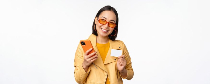 Online shopping and delivery concept. Happy korean girl in stylish clothes, holding credit card and smartphone, laughing and smiling, standing over white background.