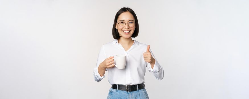 Smiling asian girl holding mug, white cup and thumbs up, recommending drink, coffee or tea, standing over white background.
