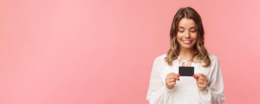 Close-up portrait of excited and amused blond girl in white dress, holding credit card and smiling thrilled, cant resist temptation to buy something, waste money online shopping, pink background.