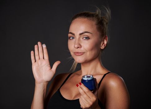 Stunning middle aged Caucasian woman with fresh glowing clear skin smiles looking at camera and holding bottle with anti-aging moisturizer facial cream, isolated over black background with copy space