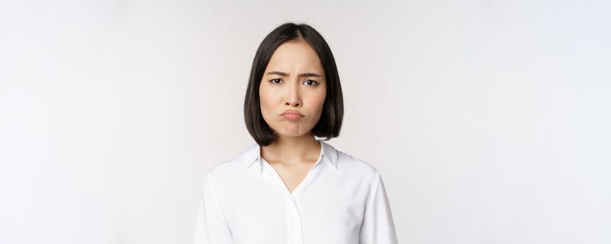 Sad and gloomy young asian woman grimacing, frowning upset, making pouting face, white background. Copy space