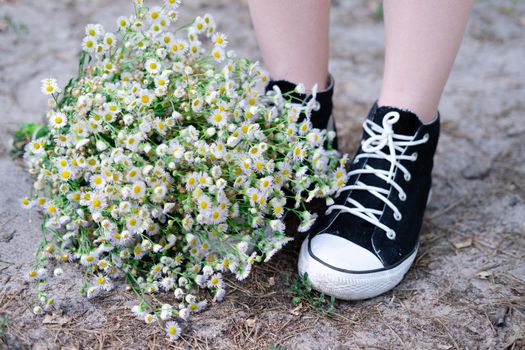 unrecognizable womens legs in sneakers near and white wildflowers outdoors on nature background on summertime.