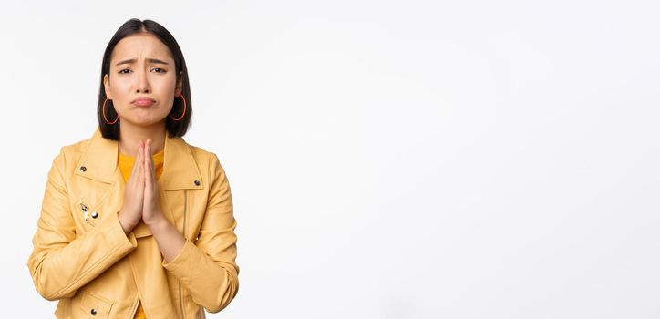 Image of hopeless asian woman begging, pleading, say sorry, looking distressed and miserable, standing over white background.