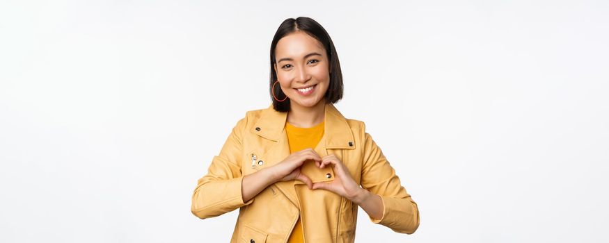 Image of beautiful asian girl showing heart, love sign, express care and romantic feelings, standing over white studio background.
