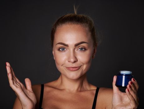 Beautiful young good looking woman with fresh glowing clear skin smiles looking at camera and holding a bottle with anti-aging moisturizer facial cream, isolated over black background with copy space