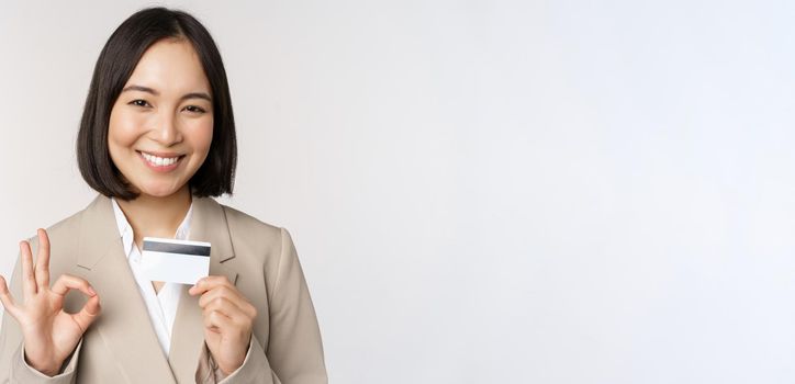 Smiling office clerk, asian corporate woman showing credit card and okay sign, recommending bank, standing over white background in beige suit.