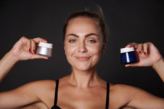 Attractive middle aged blonde European woman holds jars with anti-aging anti-wrinkles smoothing facial cream , moisturizer, smiles at camera, isolated over black background with copy ad space