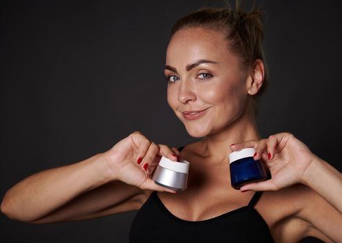 Attractive middle aged blonde Caucasian woman holds jars with anti-aging anti-wrinkles smoothing facial cream , moisturizer, smiles at camera, isolated over black background with copy ad space