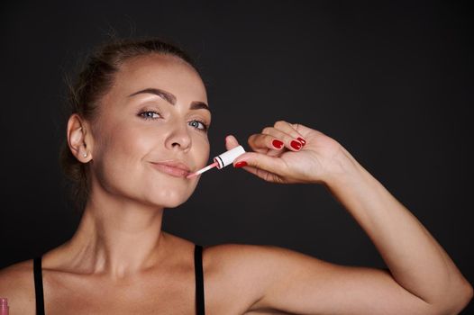 Beautiful woman applying lip gloss on her lips. Attractive blonde woman with moisturizing smoothing lip balm posing on black background with advertising copy space