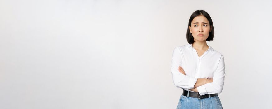Image of sad office girl, asian woman sulking and frowning disappointed, standing upset and distressed against white background.