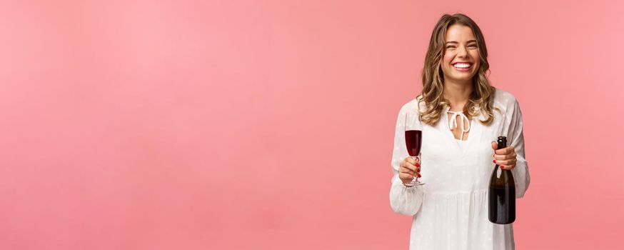 Holidays, spring and party concept. Portrait of happy and carefree european blond female celebrating in white dress, holding bottle champagne or wine, drinking from glass and laughing.