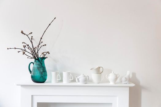 American maple branches in a green glass jar, porcelain cups, figurines, teapot on a white chest of drawers. Scandinavian style