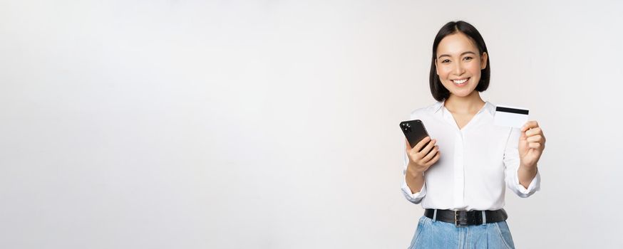Online shopping concept. Smiling modern asian girl shows her credit card, holds mobile phone, order with smartphone, standing over white background.