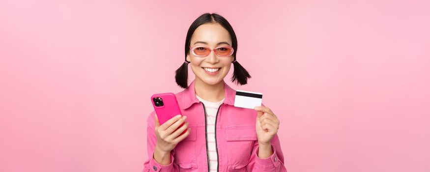 Online shopping. Smiling asian girl shopper, holding smartphone and credit card, paying in mobile app, standing over pink background.