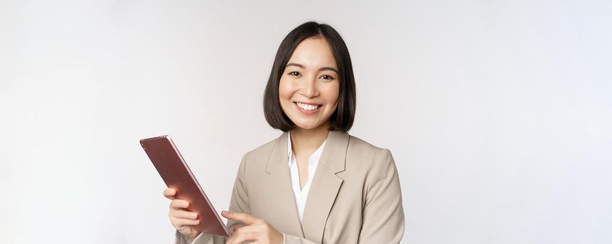 Image of asian businesswoman, saleswoman holding digital tablet and smiling, working with gadget, standing in suit over white background.